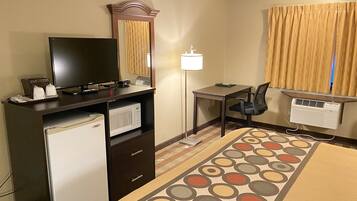 Habitación de hotel con escritorio, silla, televisor, mini refrigerador y una alfombra estampada.