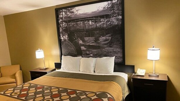 Habitación de hotel con una cama, dos mesitas de noche, una silla y un cuadro grande en la pared.
