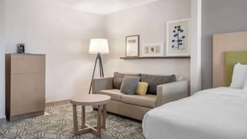 A hotel room with a bed, a sofa, a small round table, a lamp, a cabinet, and framed artwork on the wall.