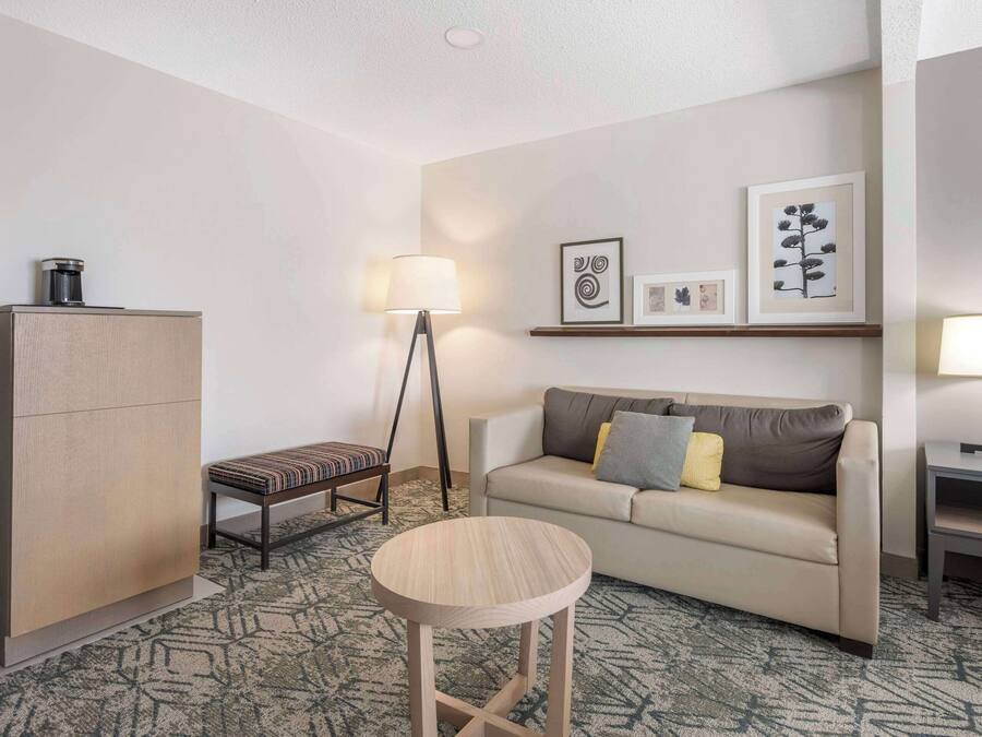 Una habitación de hotel moderna con sofá, una mesa redonda de madera, un banco, un armario y una lámpara de pie.