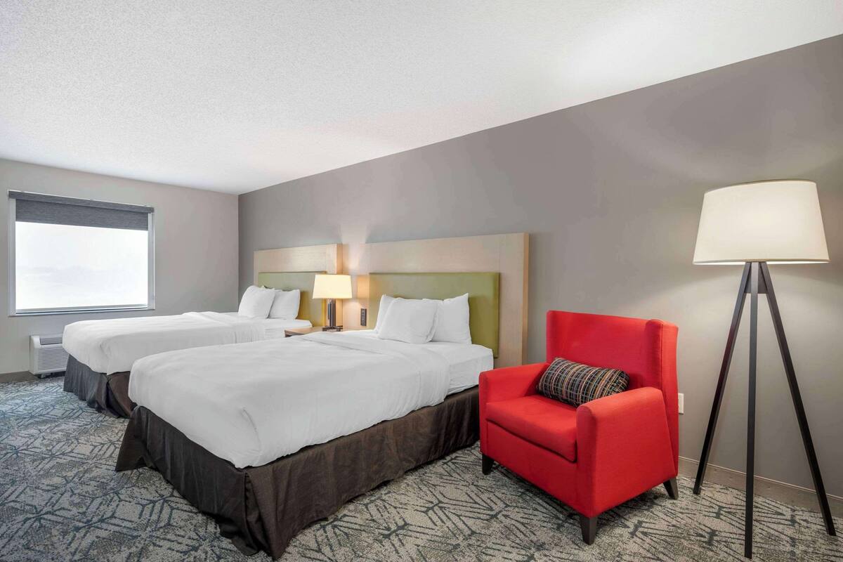 A hotel room with two beds, a red armchair, a floor lamp, and a window with blinds.