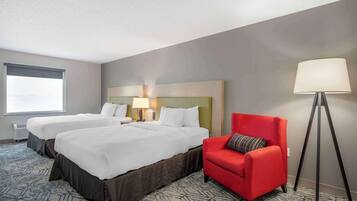 A hotel room with two beds, a red armchair, a floor lamp, and a window with blinds.