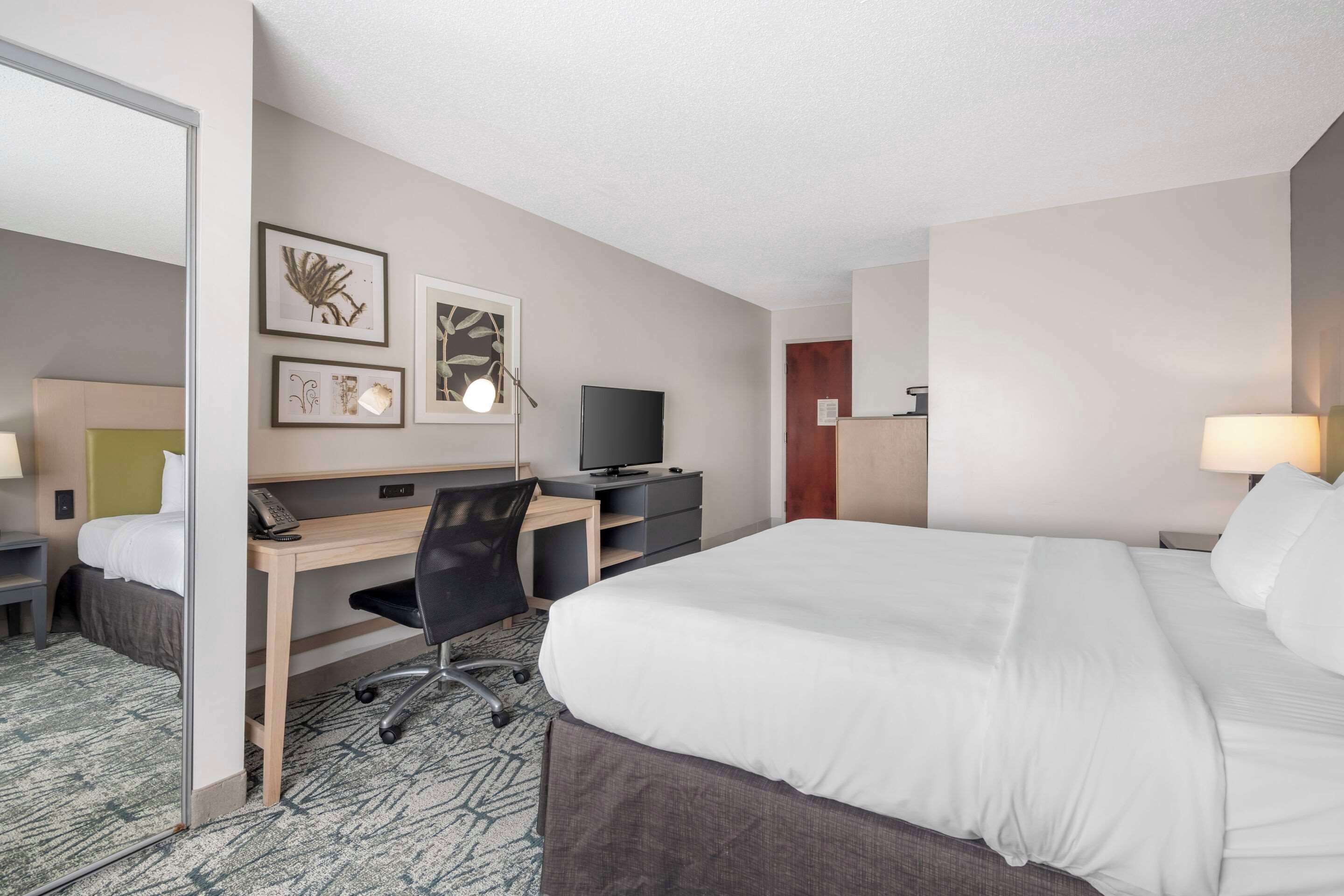 A hotel room with a bed, desk, chair, TV, and artwork on the wall.