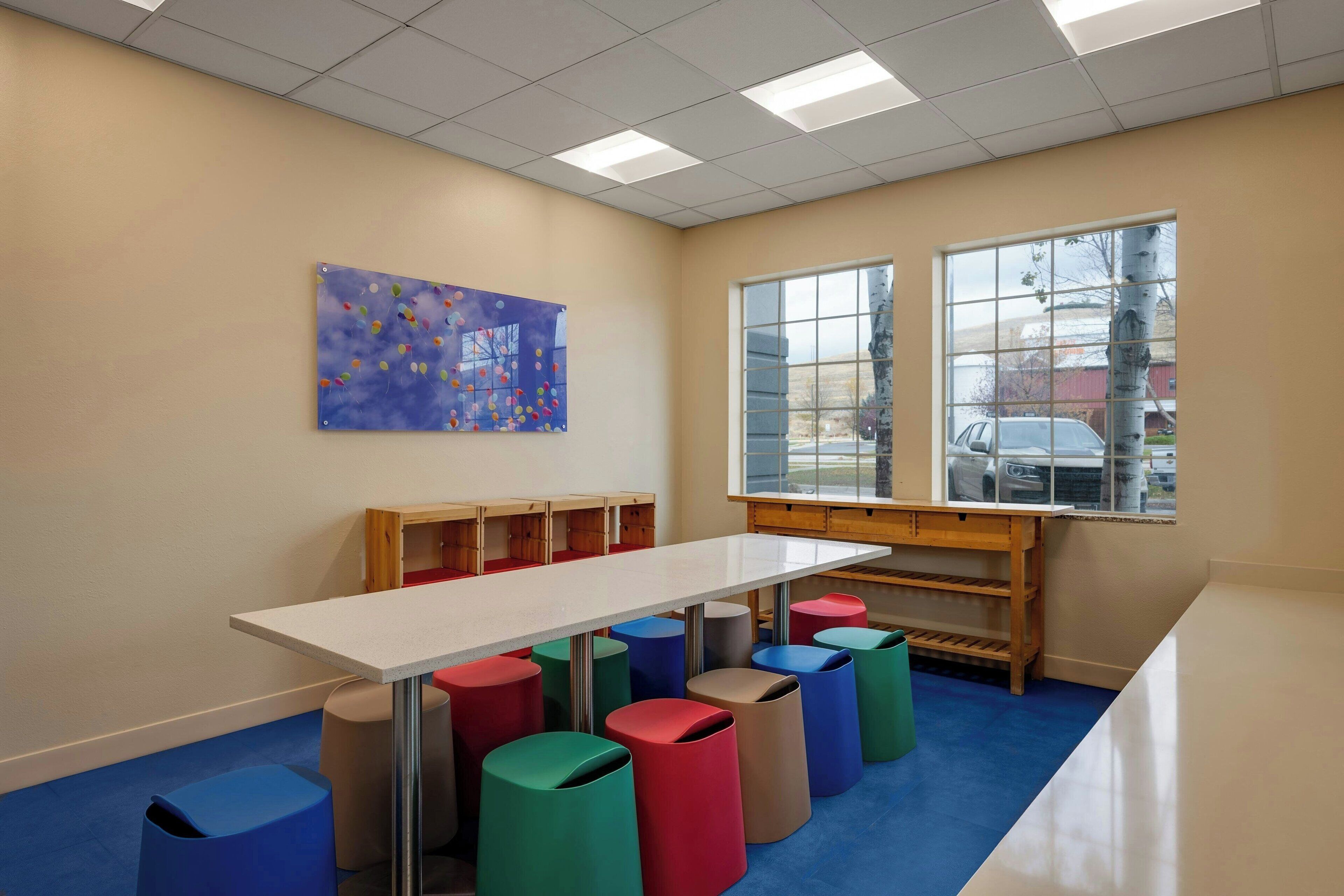 Een klaslokaal met een witte tafel, kleurrijke stoelen, een blauw kunstwerk en een raam met uitzicht op een auto en een gebouw.