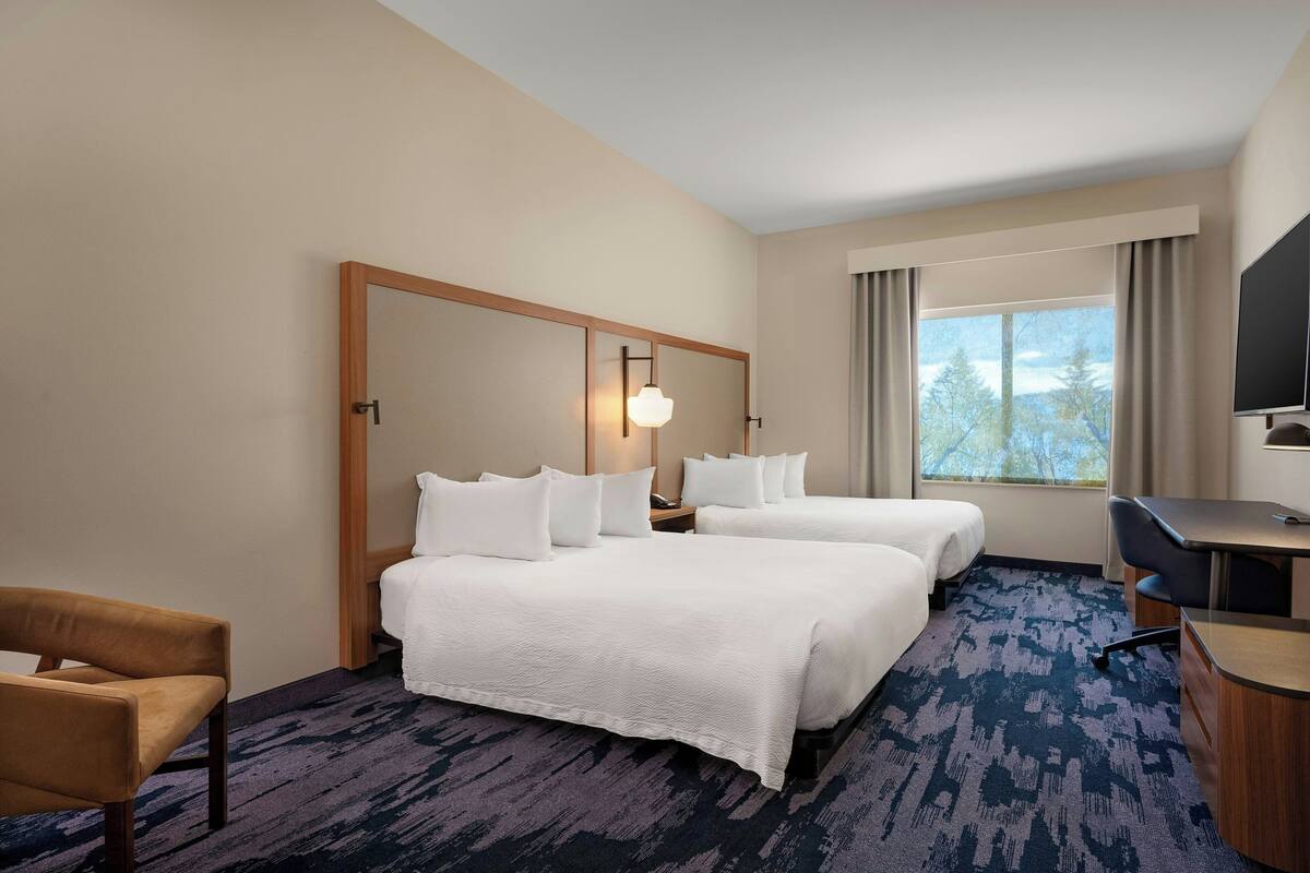 A hotel room with two beds, a desk, a chair, a TV, and a large window with a view of trees.