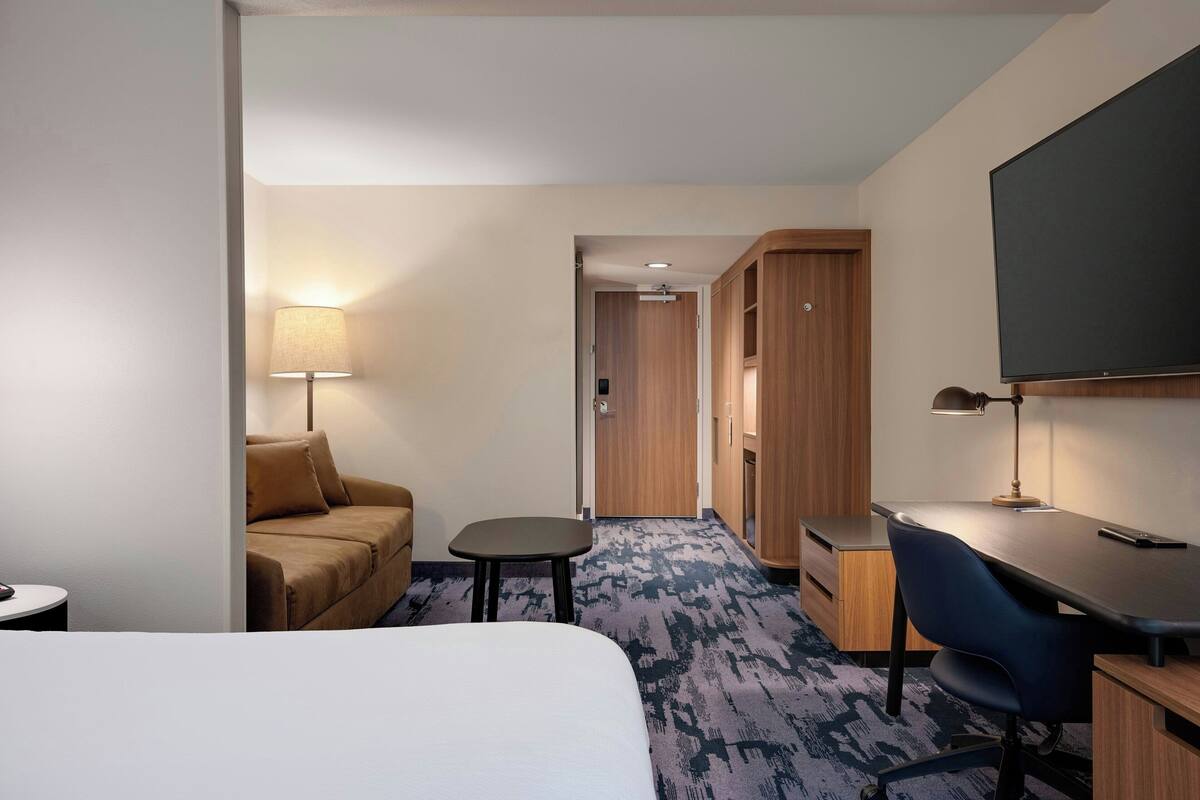 A hotel room with a bed, a desk, a chair, a small table, a lamp, a sofa, and a television.