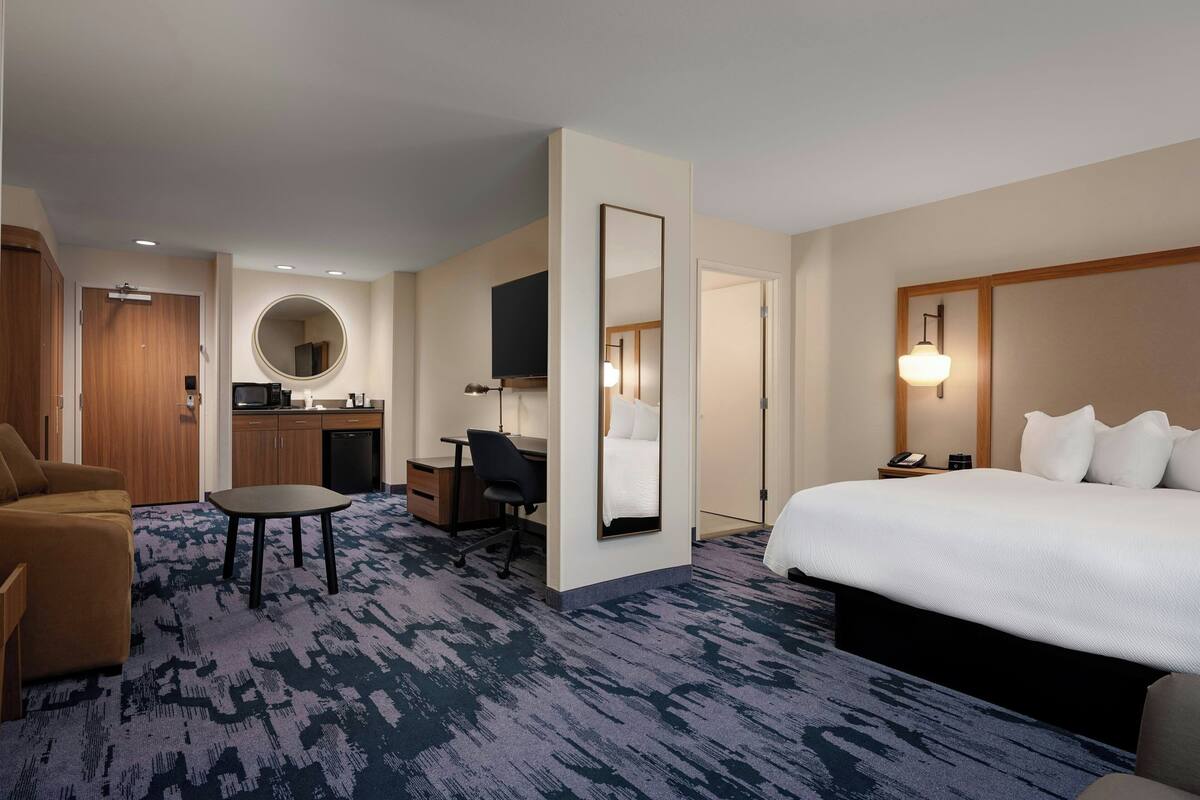 A hotel room with a bed, a desk, a chair, a small table, a mirror, and a kitchenette.