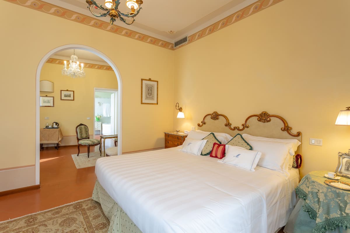 A hotel room with a large bed, a chandelier, a small table with a lamp, and a chair.
