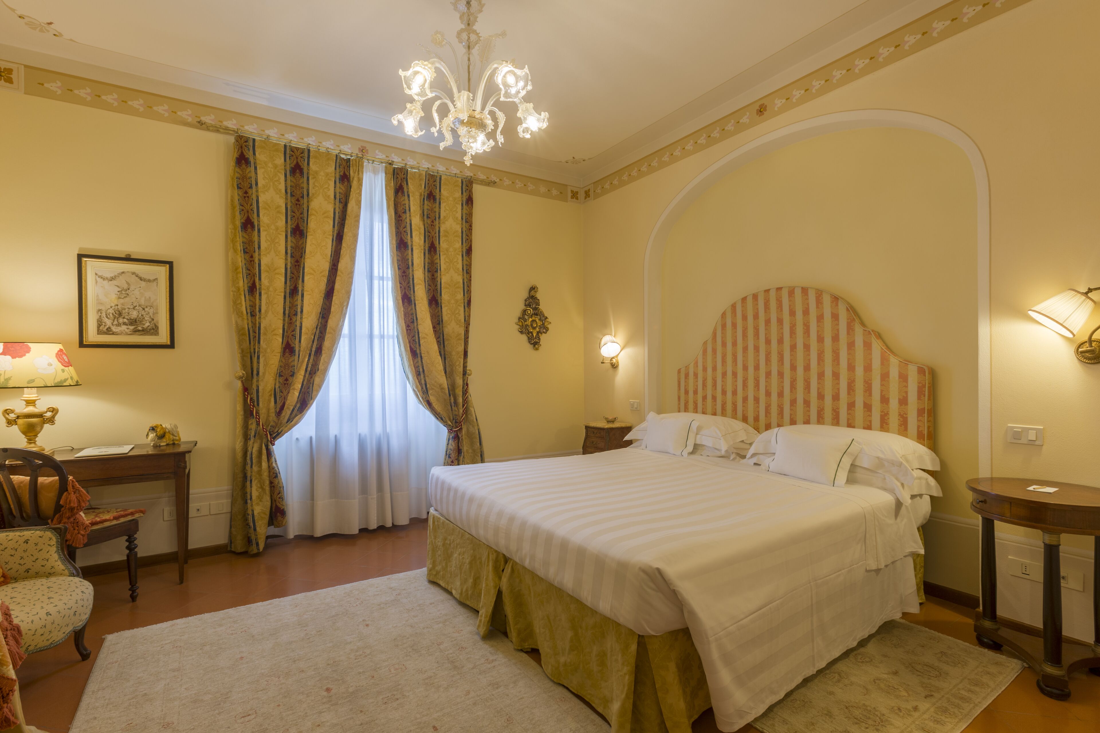 A hotel room with a large bed, a desk with a lamp, a chair, and a chandelier.