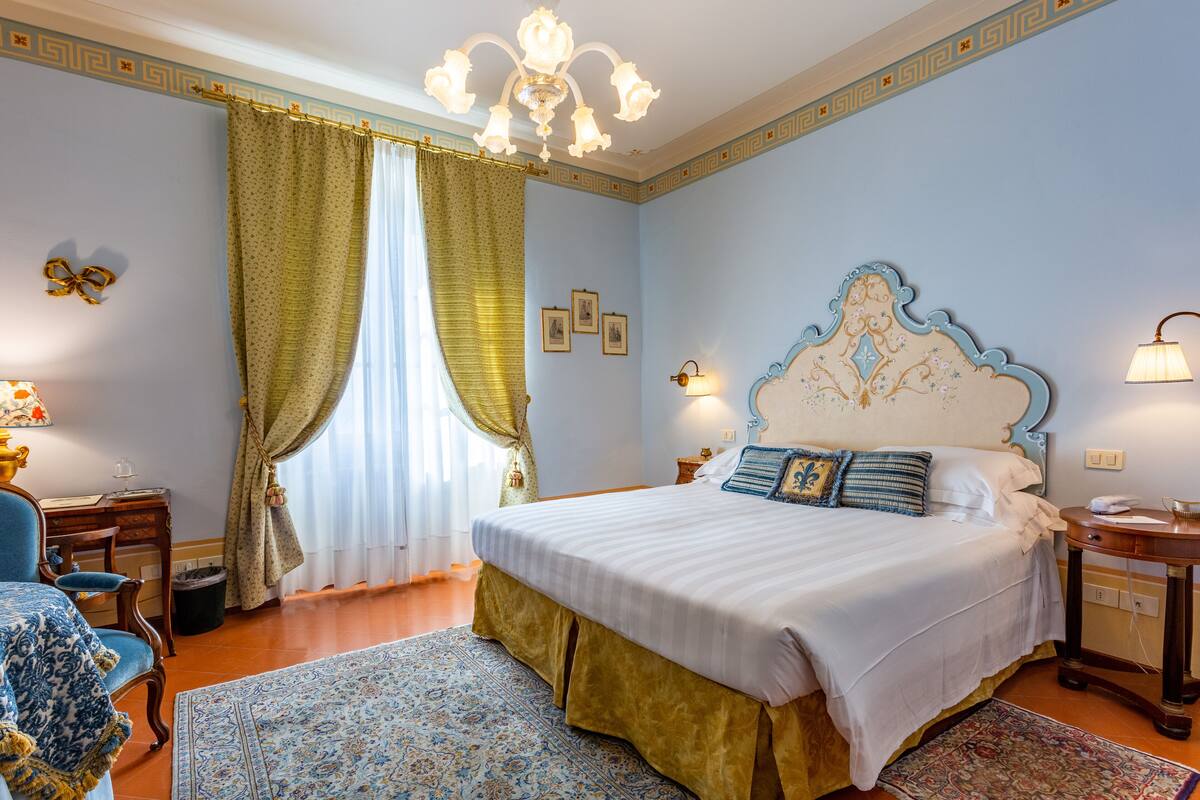 A well-lit bedroom with a large bed, a desk, and a chair. There are curtains, a chandelier, and wall-mounted lights.
