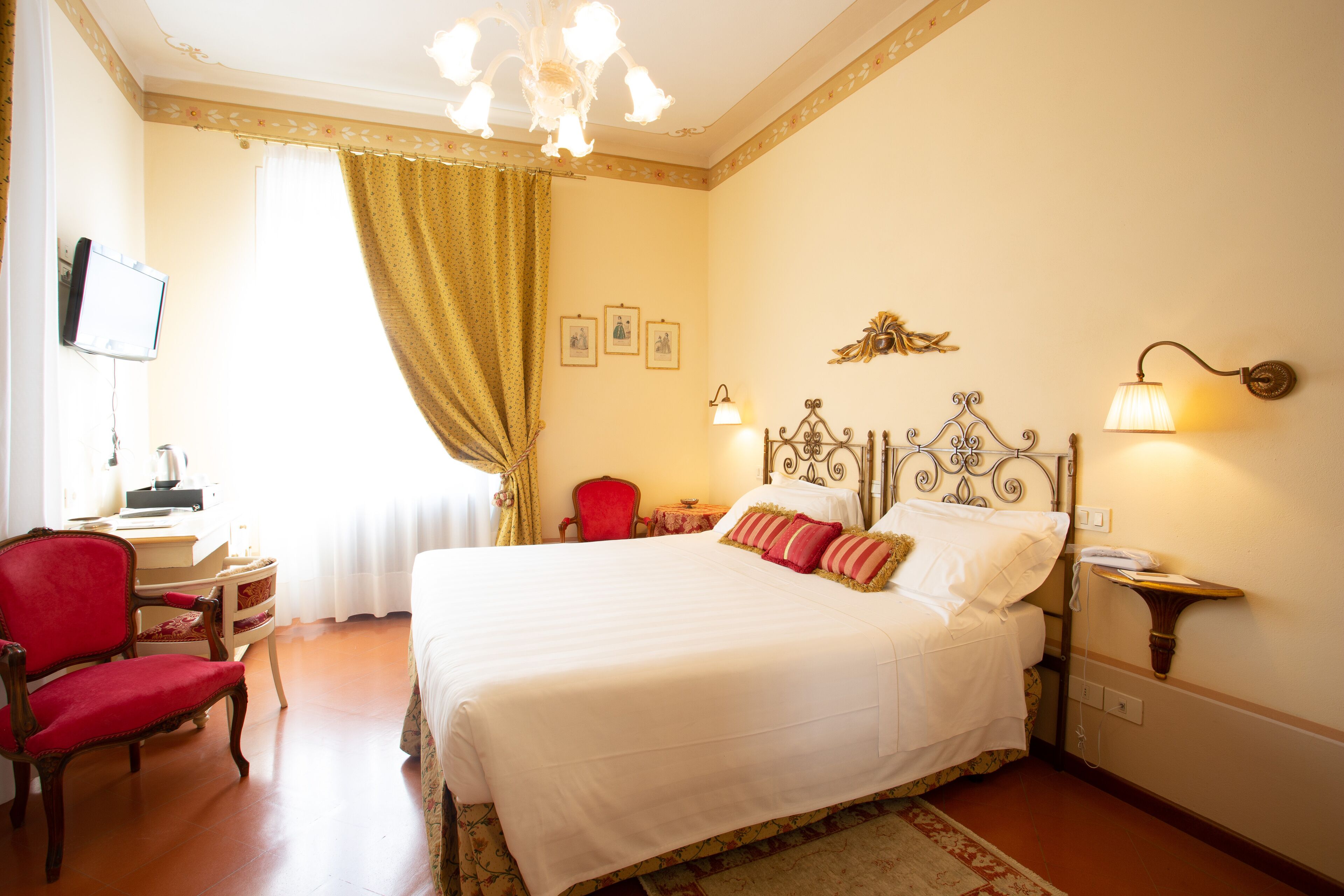A hotel room with a large bed, a red armchair, a desk with a television, and a chandelier.