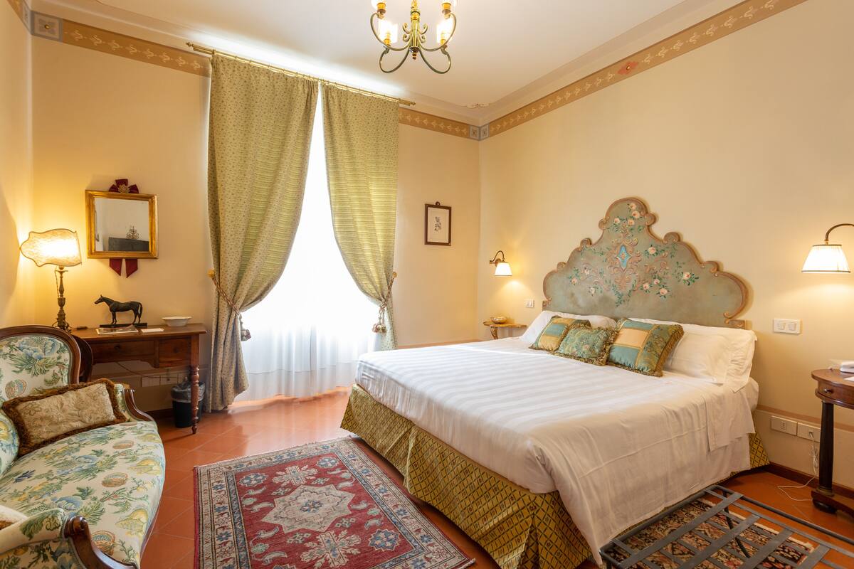 A well-lit bedroom with a large bed, a desk, and a chair. There is a window with curtains, a chandelier, and a wall-mounted lamp.