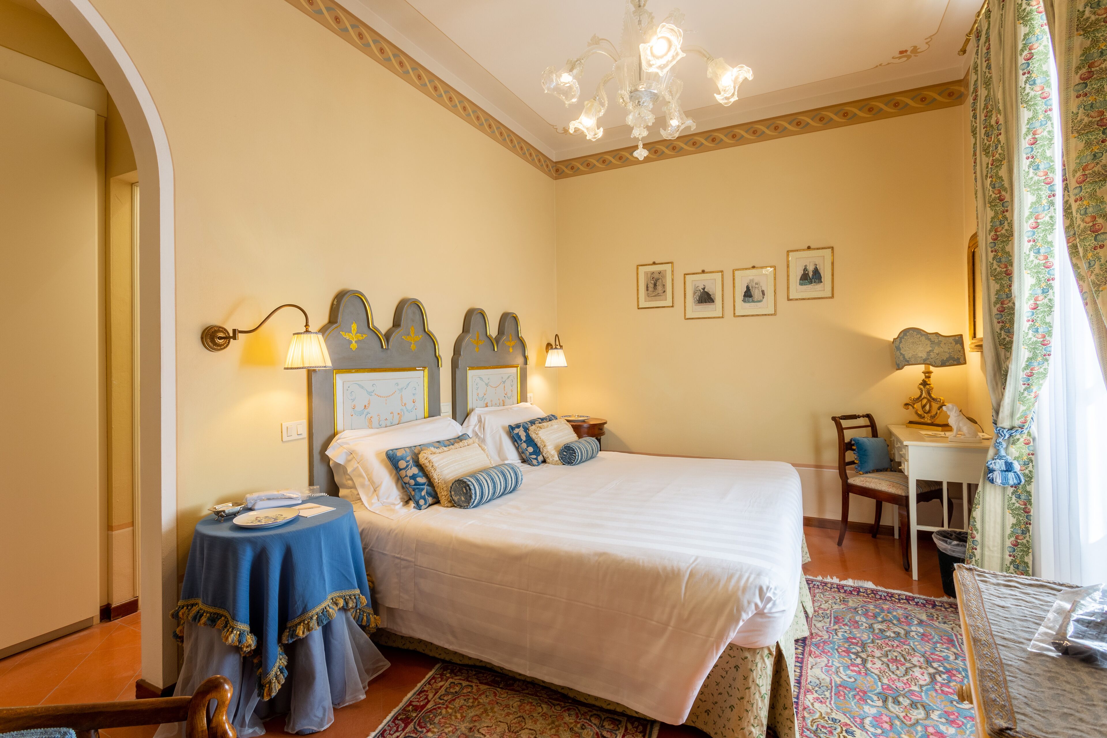 A bedroom with a large bed, a small round table, and a chair. There are wall-mounted lamps and framed pictures on the wall.