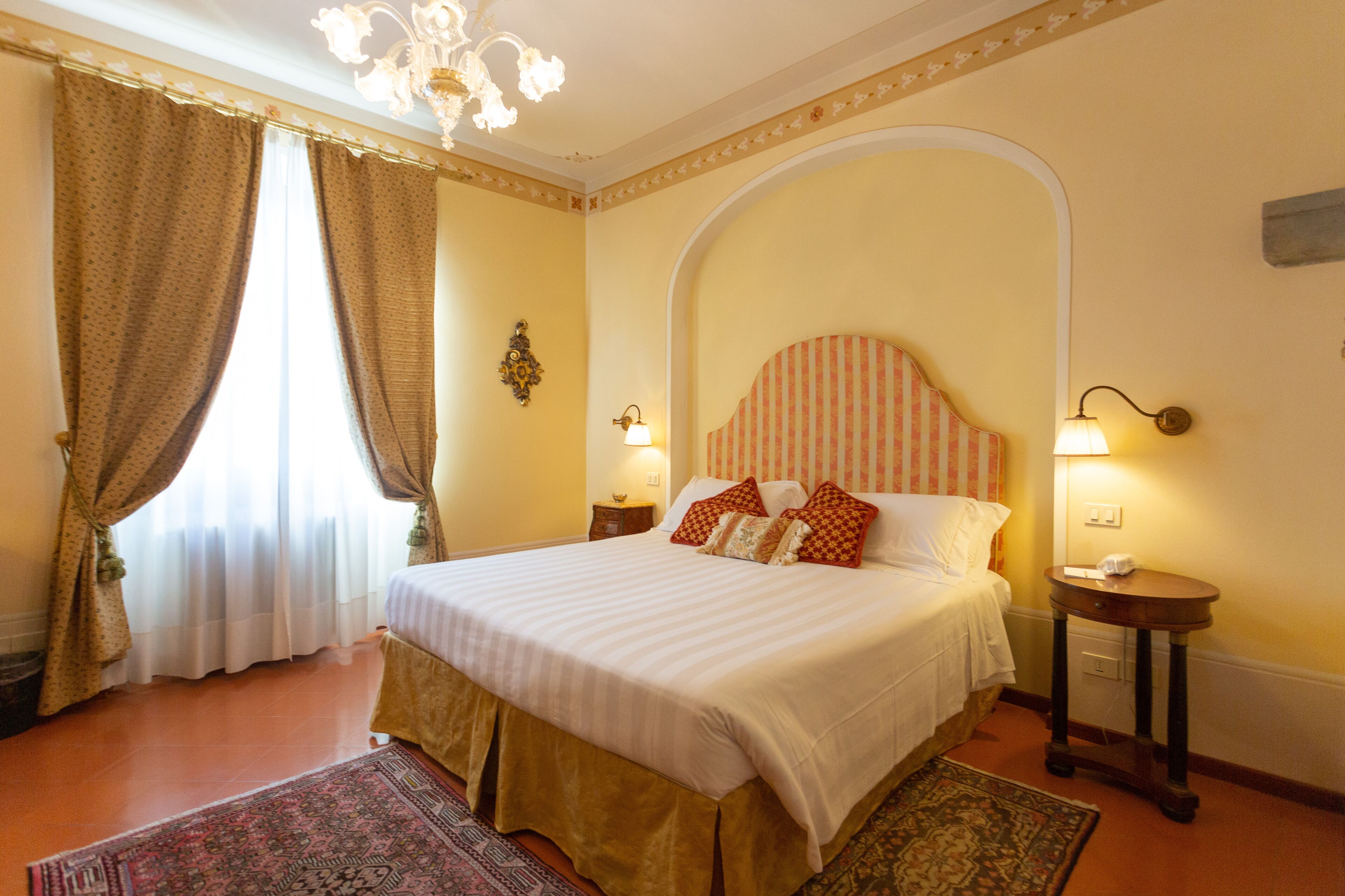 Habitación de hotel con una cama grande, dos mesitas de noche, una araña y vistas al exterior a través de cortinas.