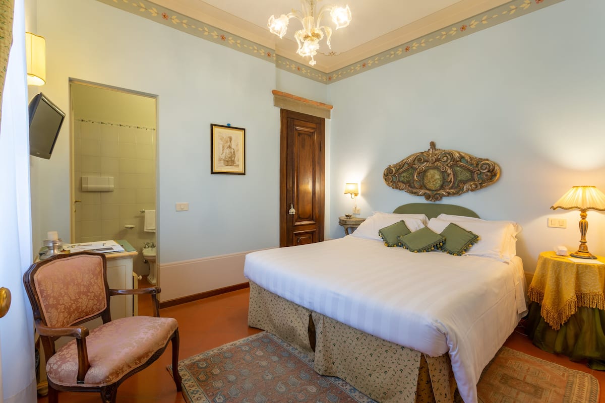 A bedroom with a bed, a chair, a desk, a mirror, and a chandelier.