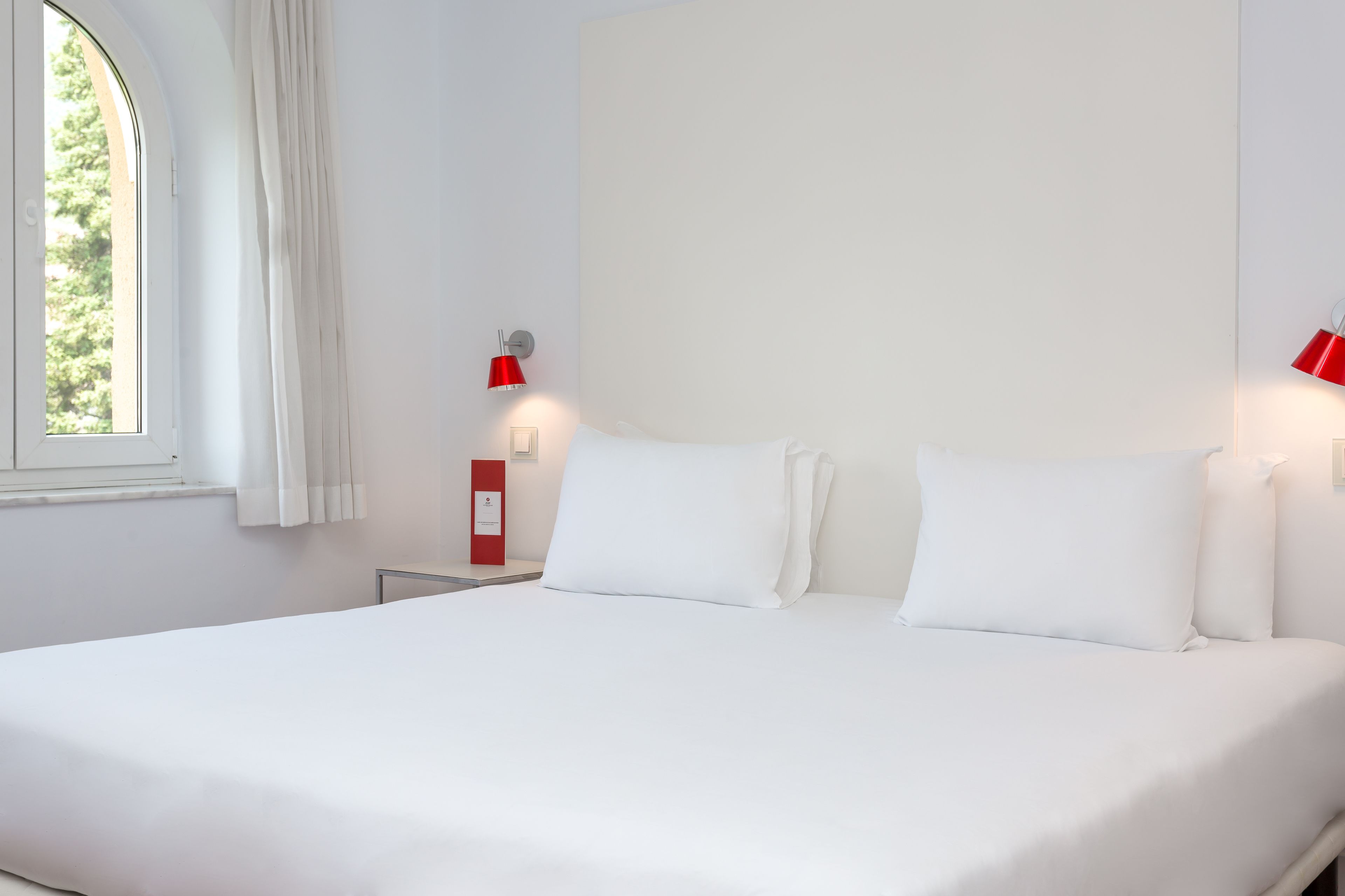 A hotel room with a large bed, white bedding, a window with curtains, and two wall-mounted red lamps.