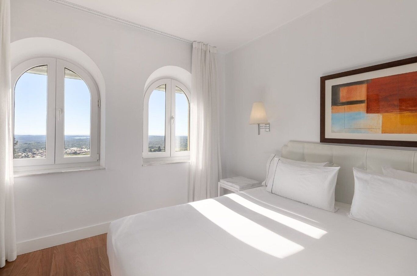 Una habitación de hotel con una cama grande, dos ventanas con cortinas, una mesita de noche, una lámpara y un cuadro en la pared.