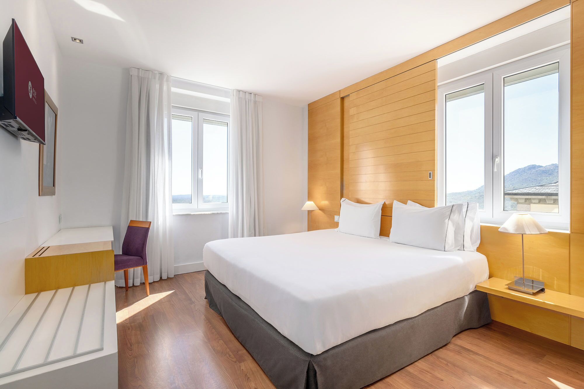 Een hotelkamer met een groot bed, een bureau, een stoel, een lamp en uitzicht op de bergen.