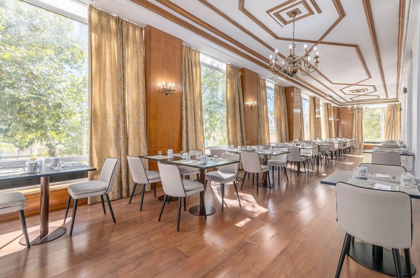 A spacious dining area with multiple tables and chairs, large windows with curtains, and a chandelier.