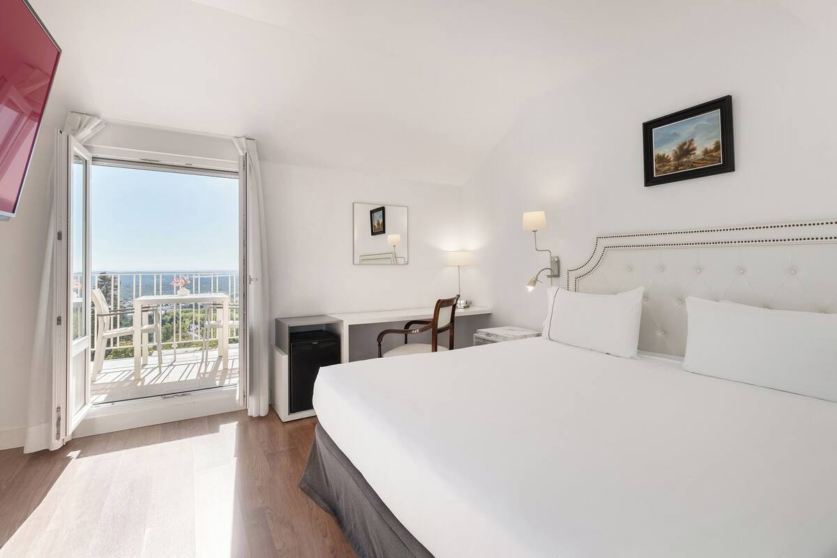 Une chambre d’hôtel avec un grand lit, un bureau, une chaise, une petite table, une lampe et un tableau au mur.