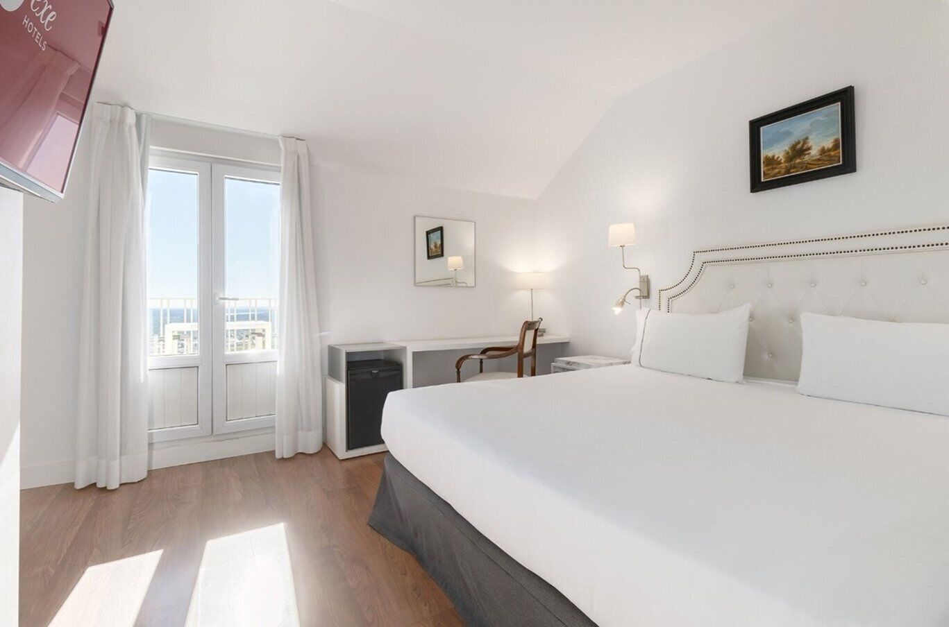 Une chambre d’hôtel avec un grand lit, un bureau, une chaise, un petit placard et une fenêtre avec des rideaux.