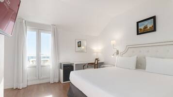 Une chambre d’hôtel avec un grand lit, un bureau, une chaise, un petit placard et une fenêtre avec des rideaux.