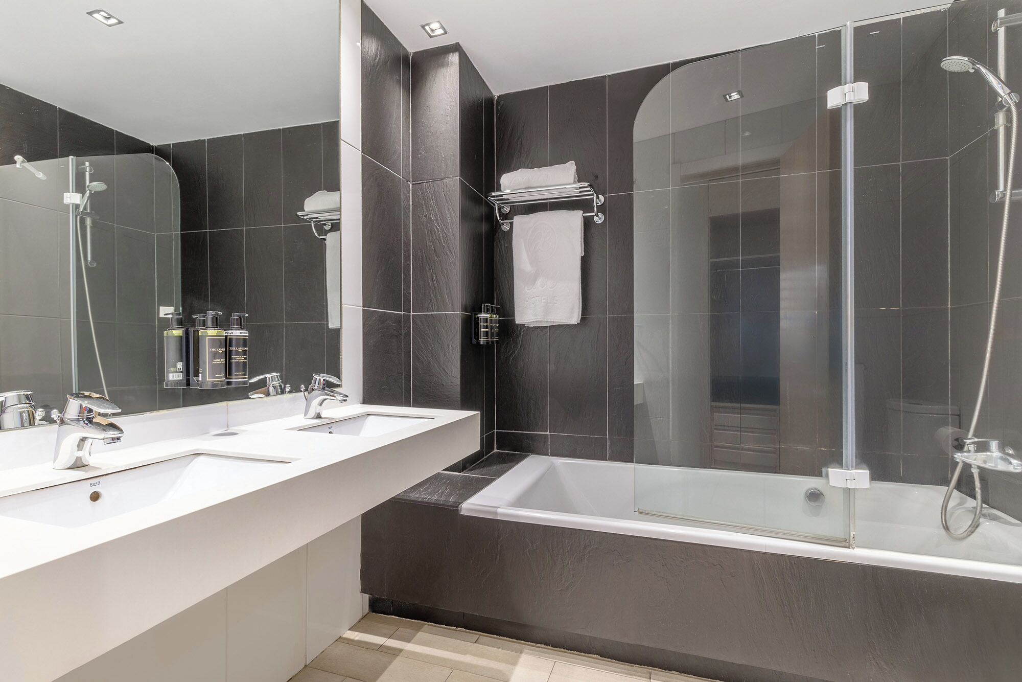 A modern bathroom with two sinks, a large mirror, a glass-enclosed shower, and dark tiled walls.