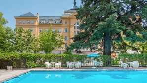 Una piscina con sillones de descanso y sombrillas, rodeada de árboles y un edificio grande con una torre.