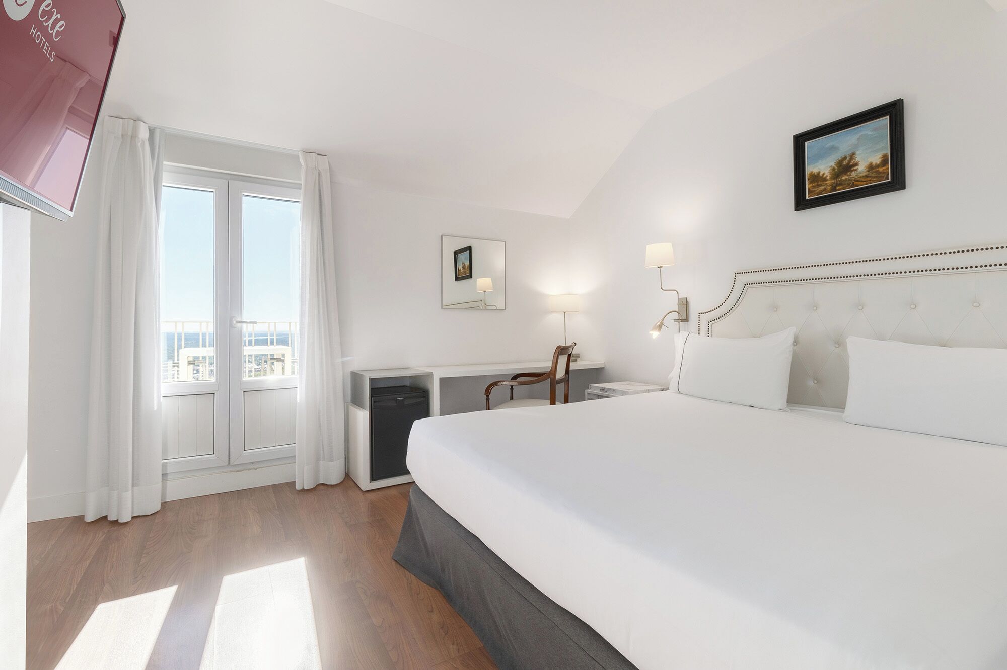 Ein Hotelzimmer mit einem großen Bett, einem Schreibtisch mit Stuhl, einer kleinen Kühlbox, einem Fenster mit Vorhängen und einem Bild an der Wand.