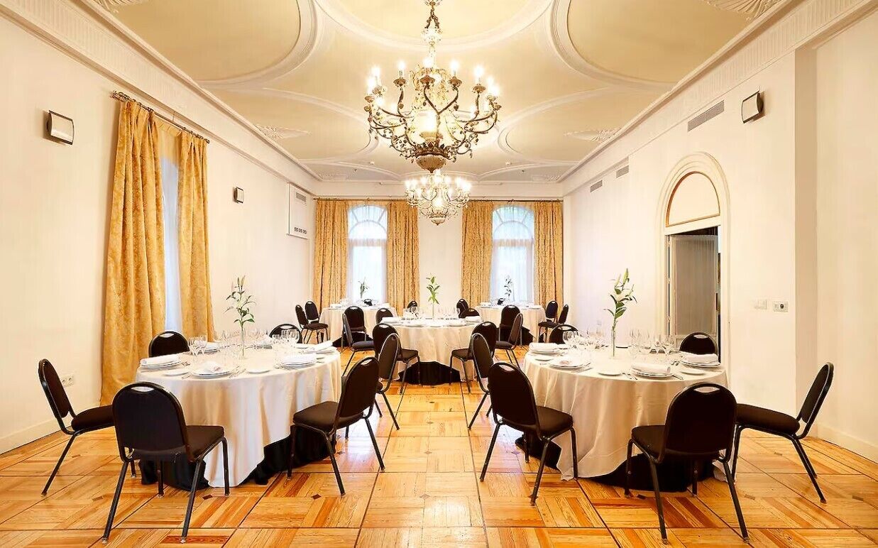 A formal dining room with round tables, black chairs, and chandeliers.