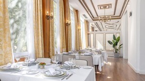 Un restaurante con varias mesas dispuestas para comer, ventanas amplias con cortinas y un candelabro.
