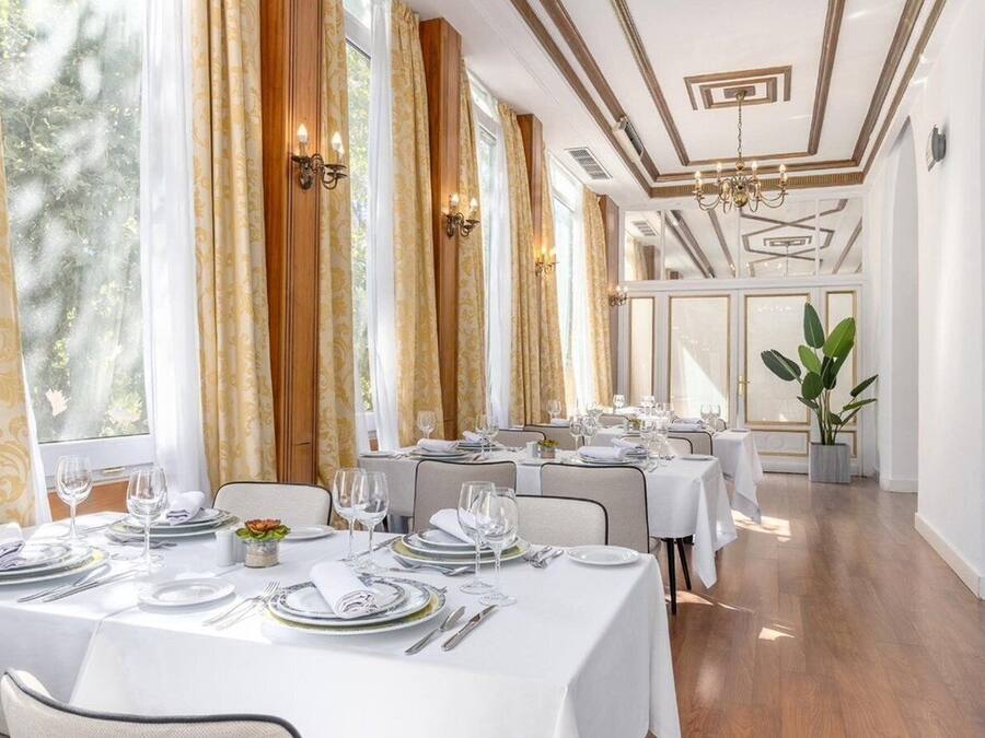 A restaurant with multiple tables set for dining, large windows with curtains, and a chandelier.