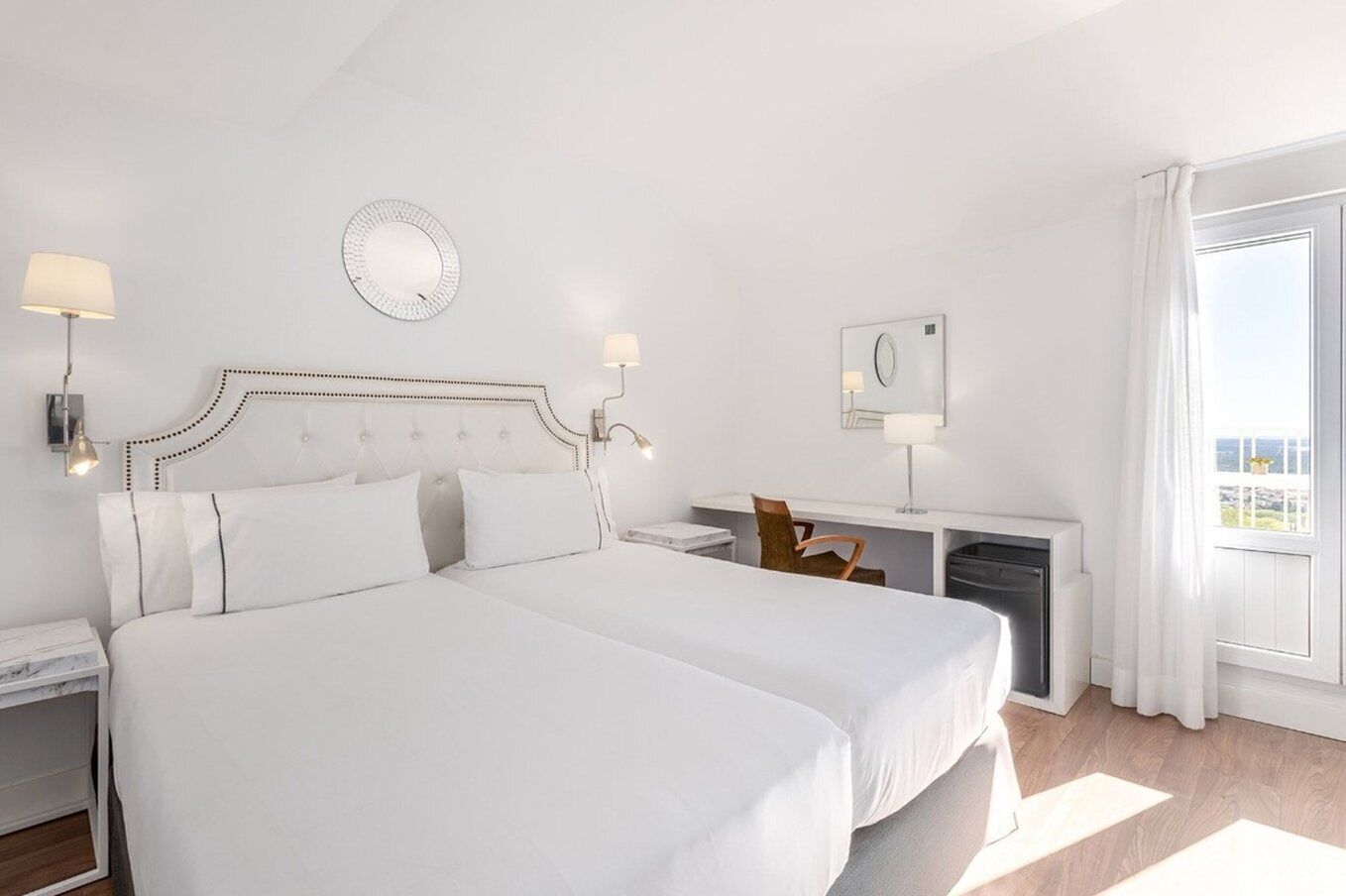 A hotel room with a large bed, a desk with a chair, a small table, a mirror, and a window with curtains.