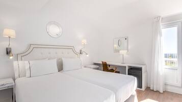 A hotel room with a large bed, a desk with a chair, a small table, a mirror, and a window with curtains.