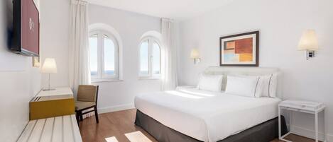 Habitación de hotel con una cama grande, dos mesitas de noche, una silla, un televisor y dos ventanas con cortinas.