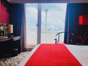 A hotel room with a bed, a chair, a desk with a lamp, and a view of a tower and cityscape through the window.