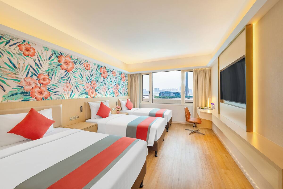 A hotel room with three beds, a large window with a city view, and a flat-screen TV.