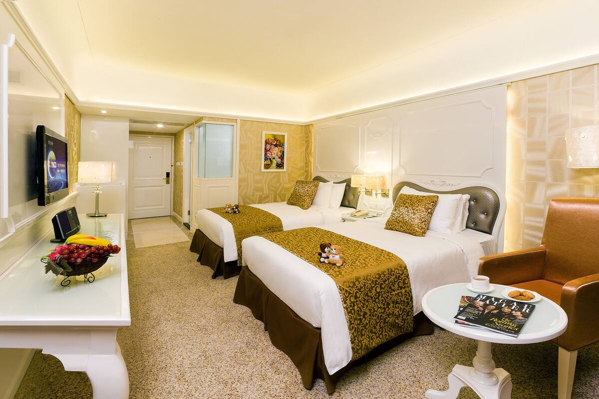A hotel room with two beds, a desk with a fruit bowl, a small table with a magazine, and a chair.