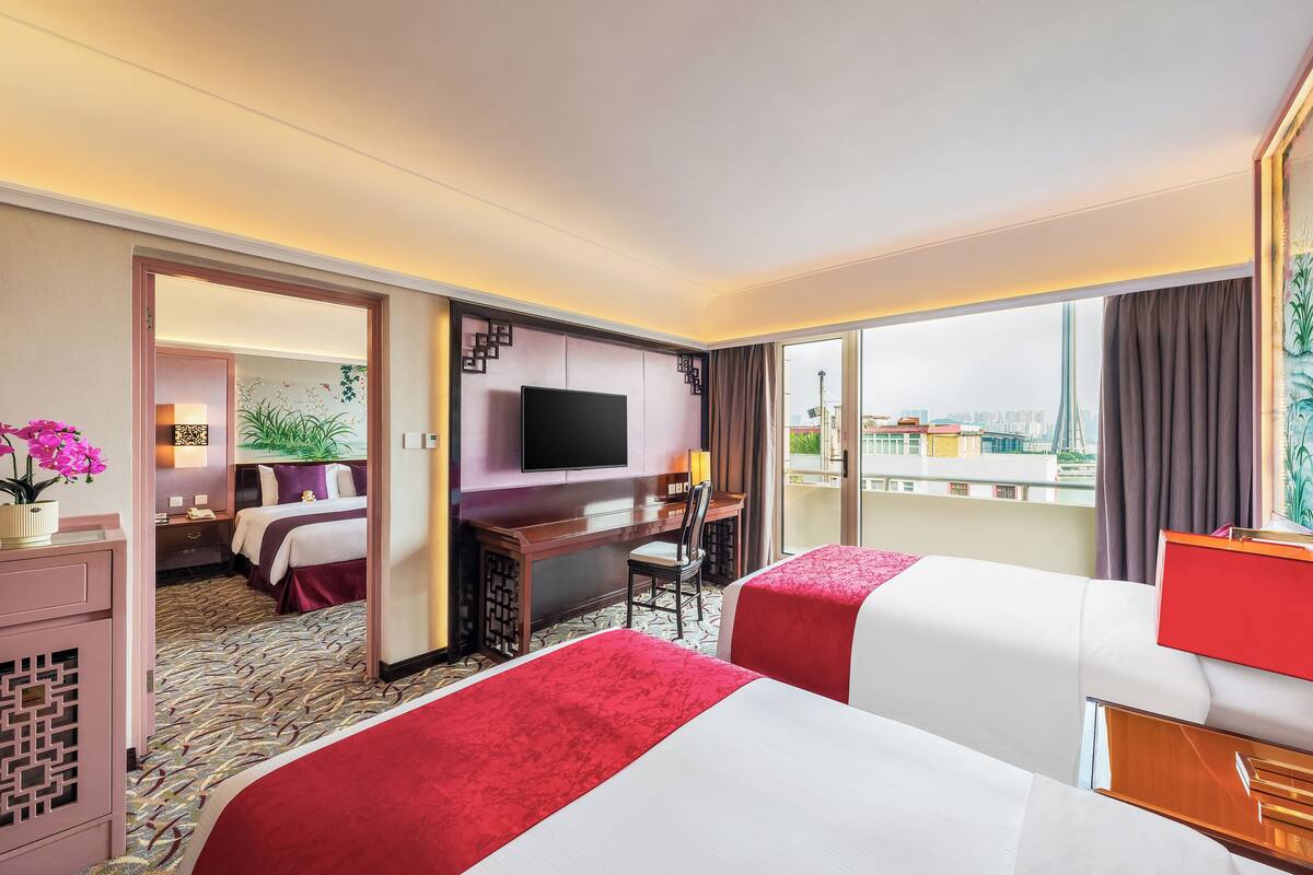 A hotel room with two beds, a desk, a TV, and a balcony with a city view.