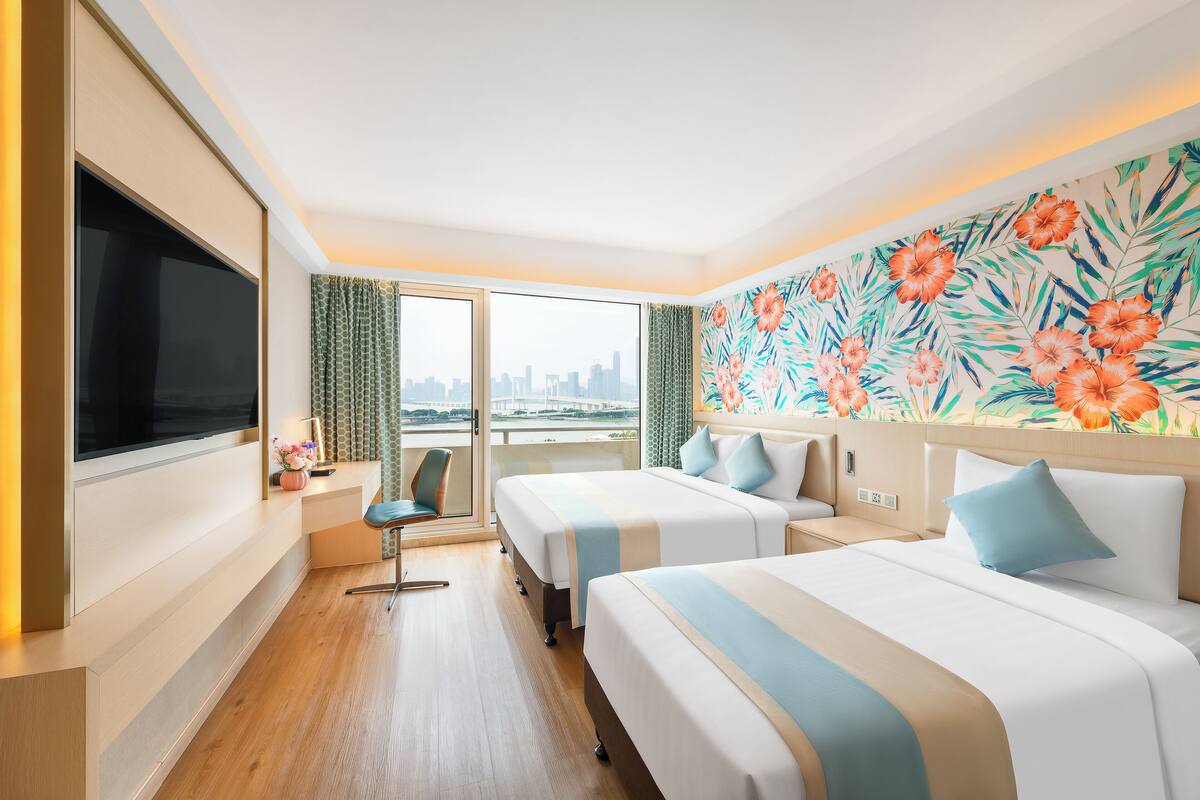 A hotel room with two beds, a TV, a desk, and a city view.