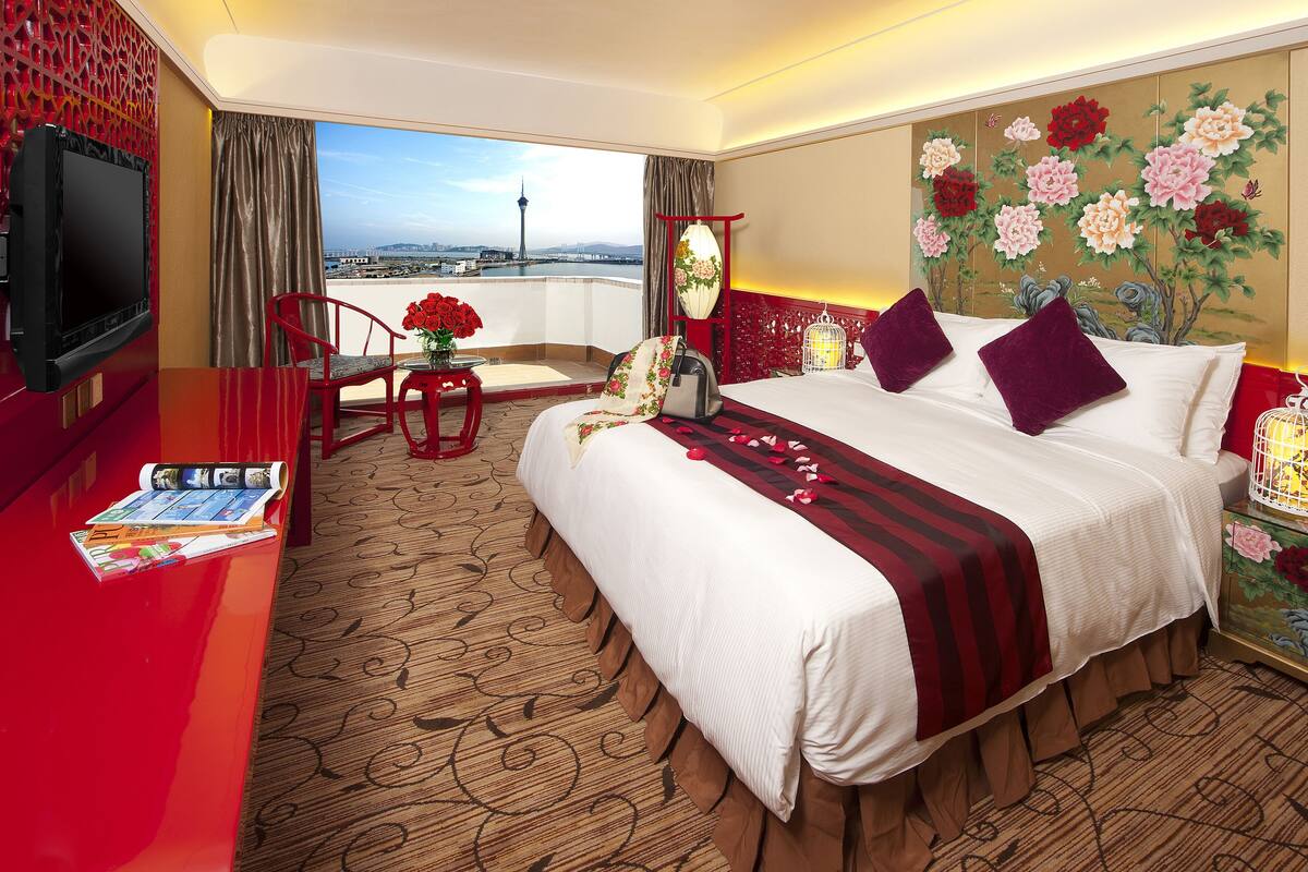 A hotel room with a large bed, a TV, a desk with a magazine, a chair, a vase of flowers, and a view of a cityscape.