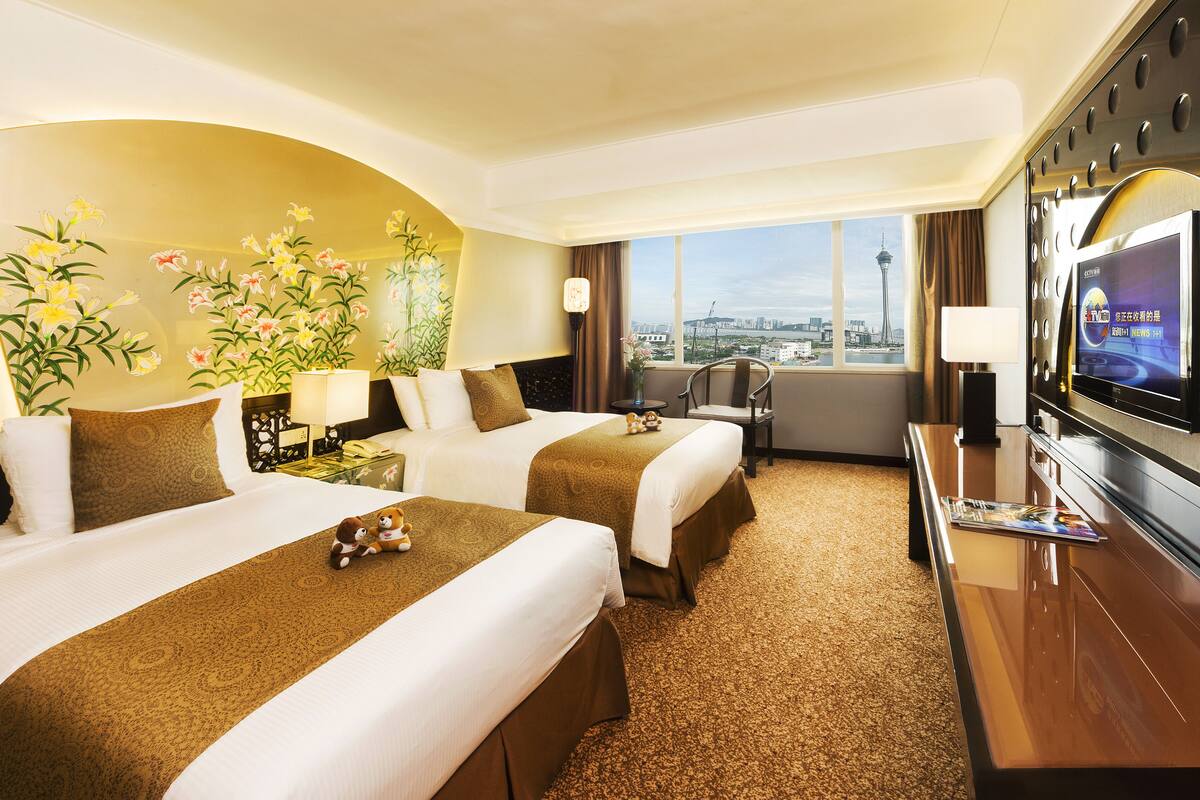 A hotel room with two beds, a TV, a desk, and a large window with a city view.