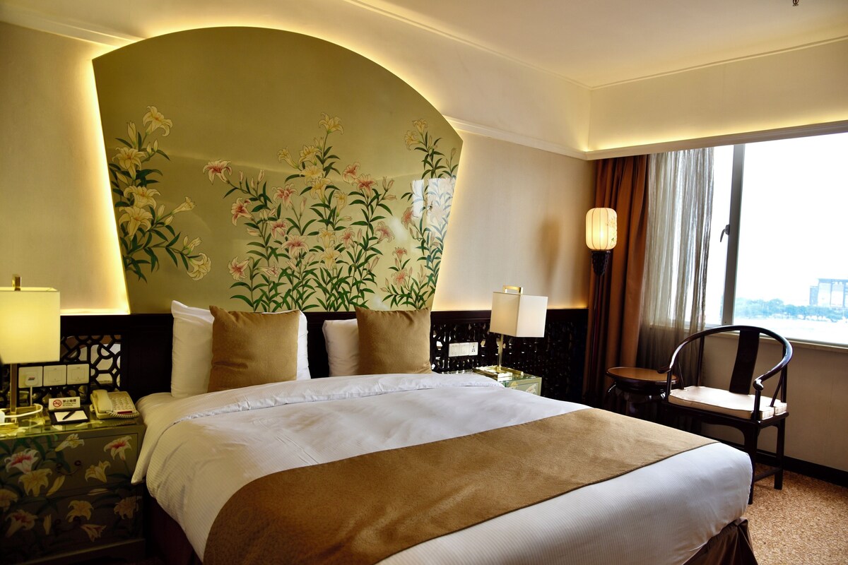 A hotel room with a large bed, a nightstand with a lamp, a chair, and a floral wall mural.