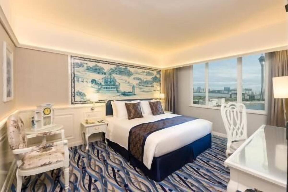 A hotel room with a bed, a desk, a chair, a large window, and a painting on the wall.