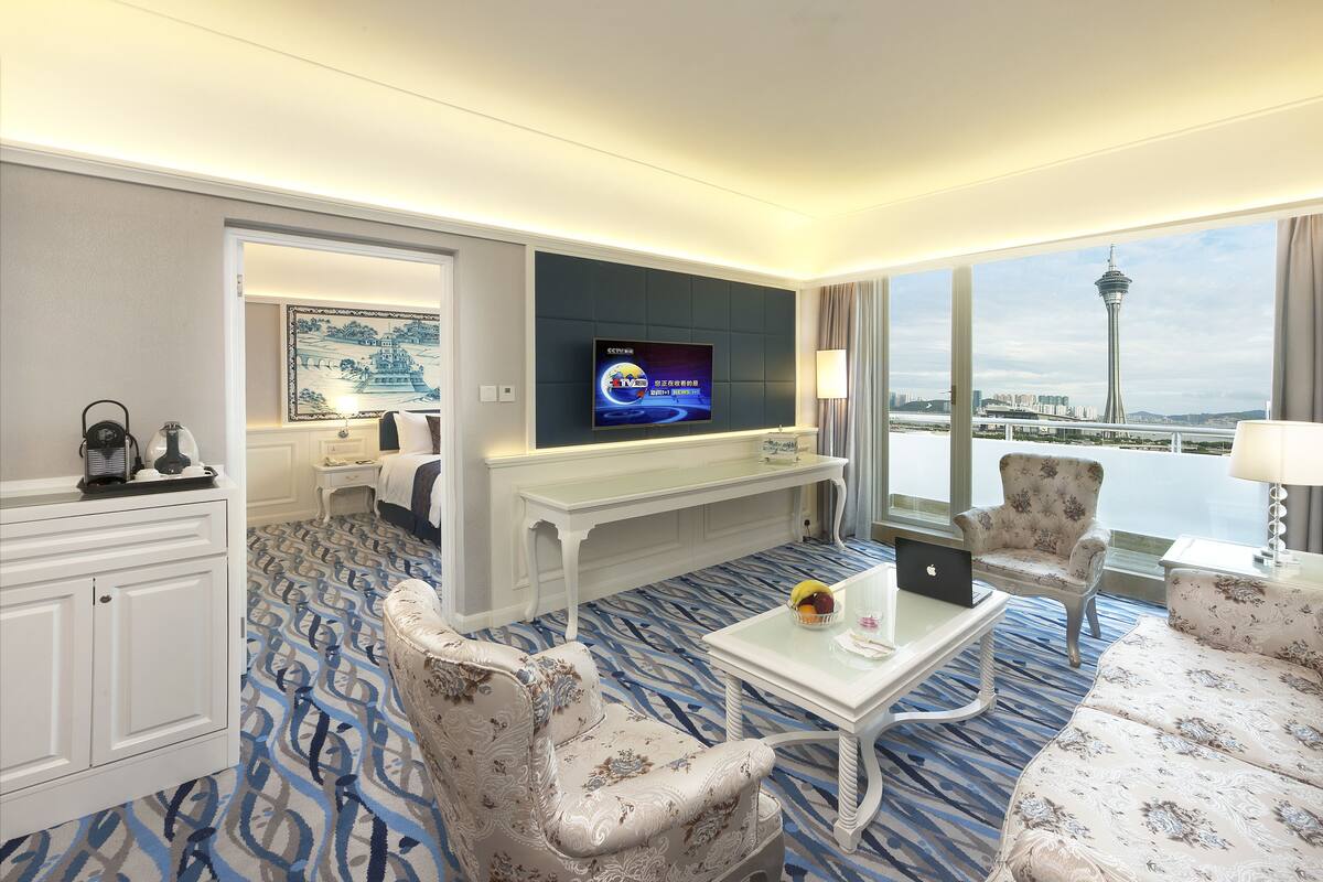 A hotel room with a large TV, a desk, two armchairs, a sofa, and a view of a tower and water.