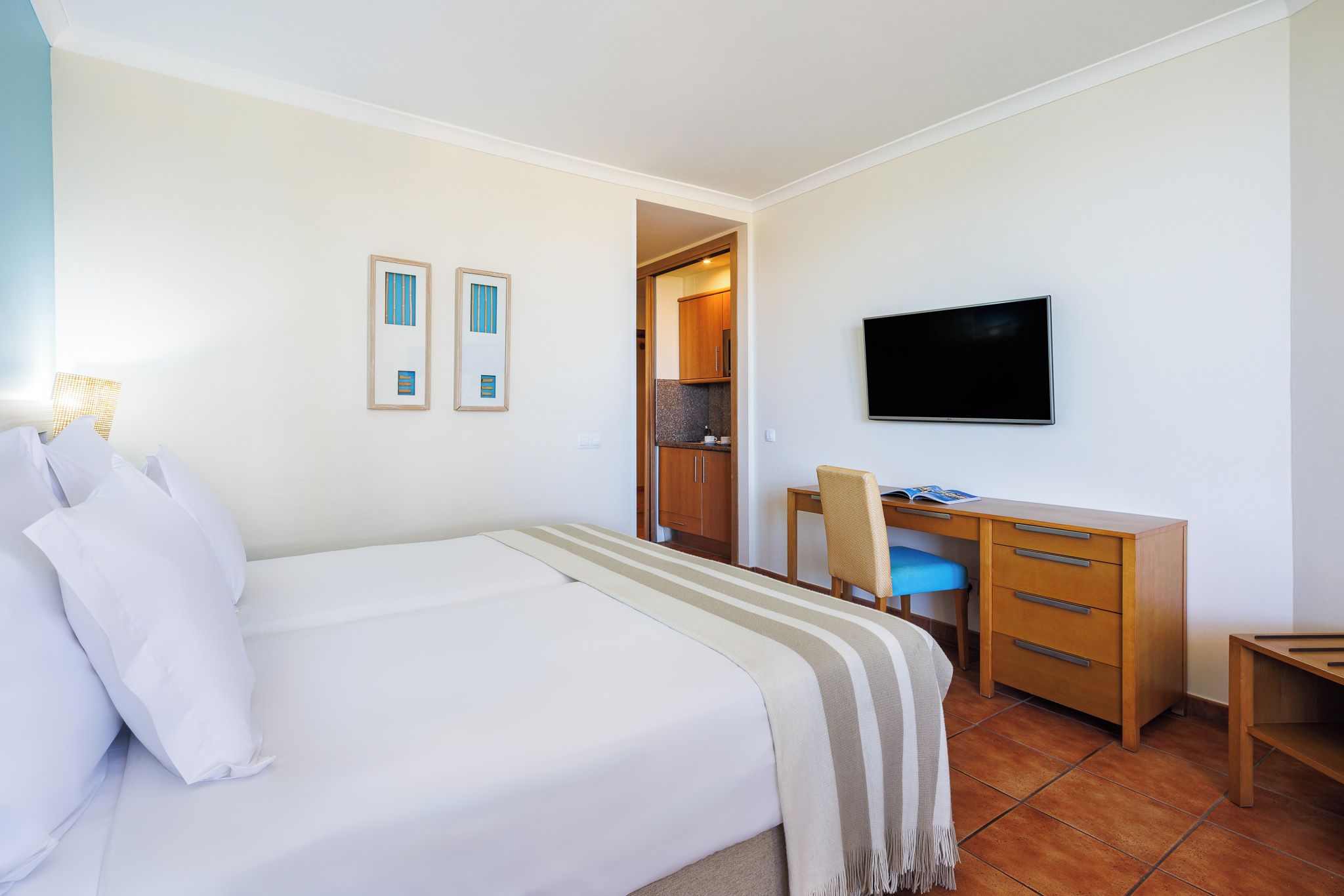 A hotel room with a bed, a desk, a chair, a television, and a door leading to a kitchenette.