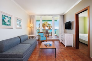 Una sala de estar con un sofá, una mesa con una revista, un televisor, un balcón con vistas a palmeras y al mar, y una ventana con cortinas.