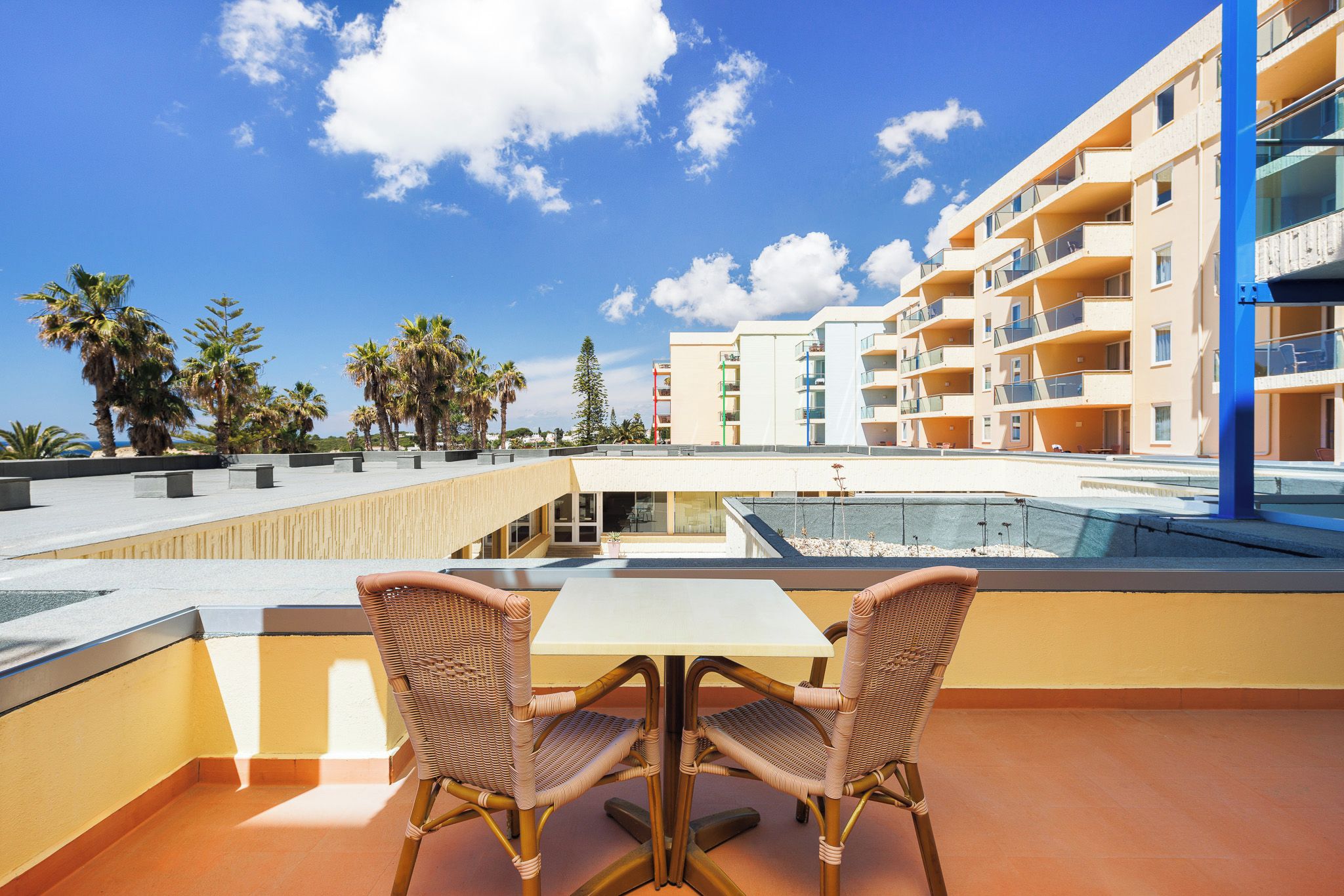 A balcony with two wicker chairs and a table, overlooking a pool and a building with multiple balconies.