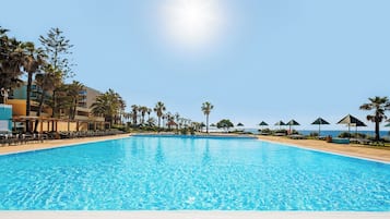 Una piscina azul cristalina con sillas de descanso y sombrillas, rodeada de palmeras y un edificio.