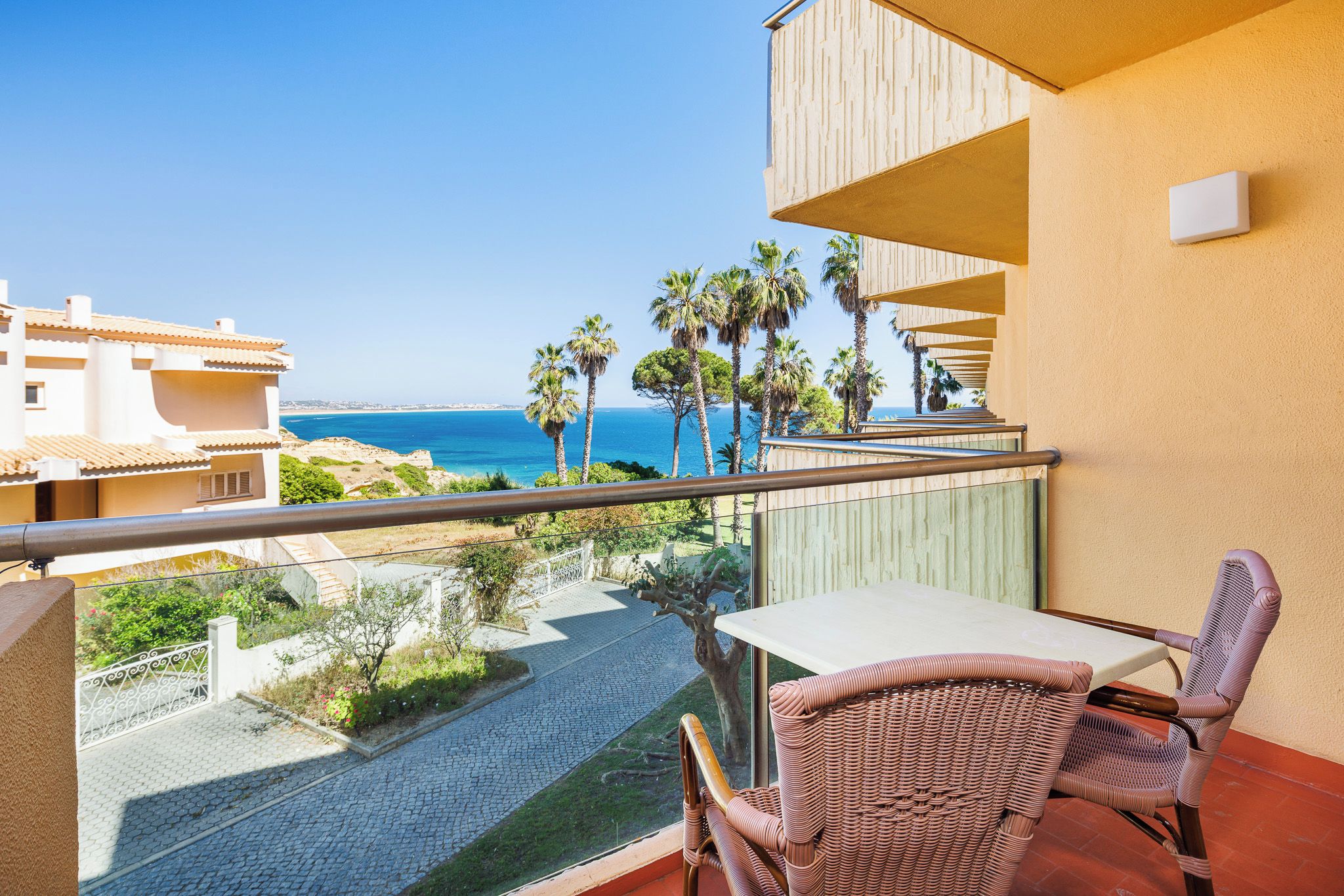 A balcony with a table and chairs, overlooking a coastal area with palm trees and a view of the sea.